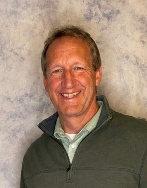 A smiling man in a casual outfit stands in front of a textured background. He's wearing a green zip-up jacket over a lighter shirt.