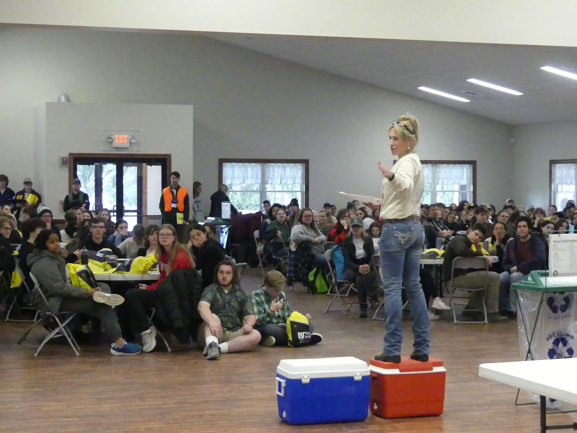 A person stands on coolers addressing a crowd seated at tables in a hall.