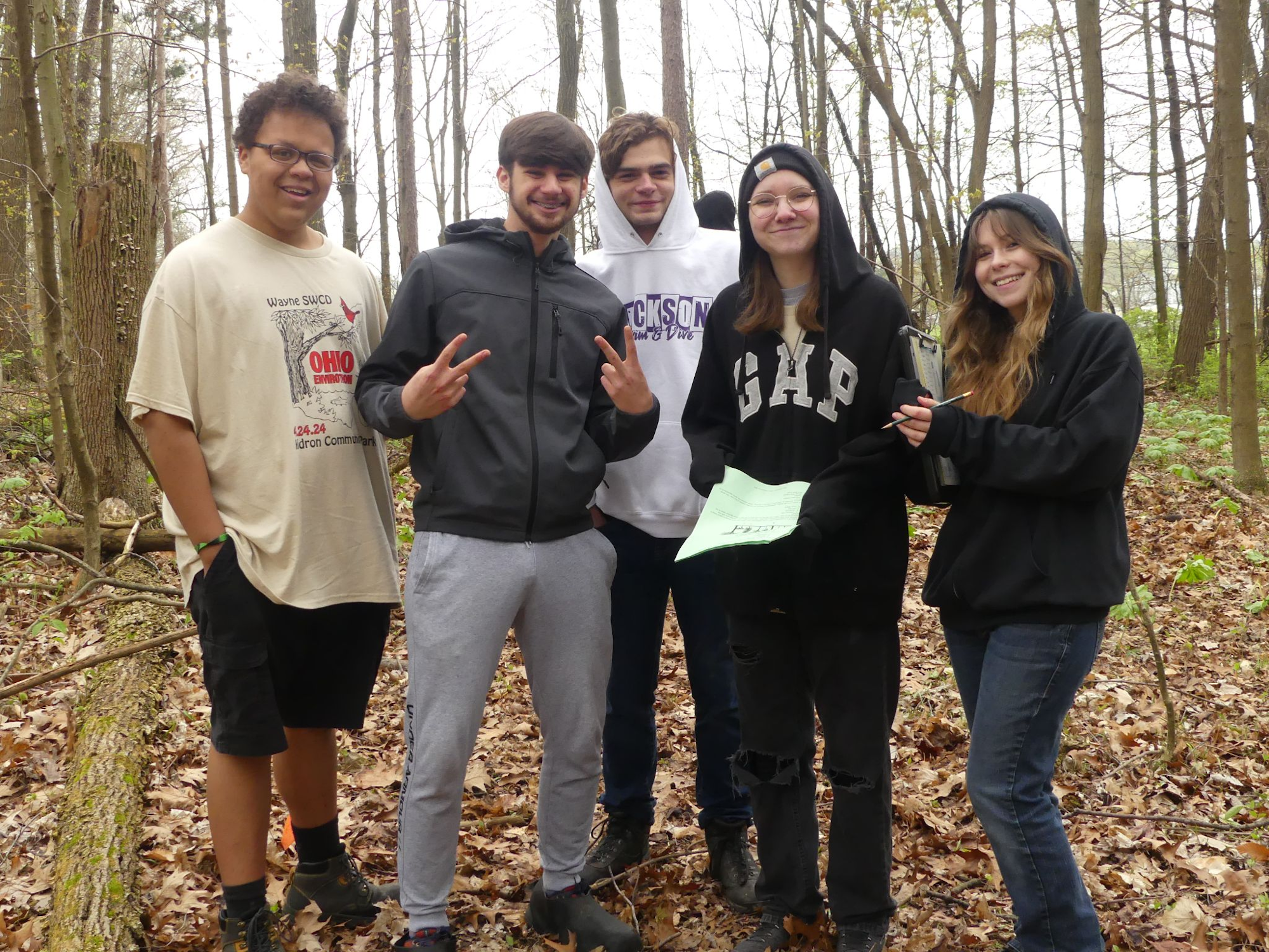 Five people smiling in a wooded area, some holding papers, wearing casual clothing and hoodies.