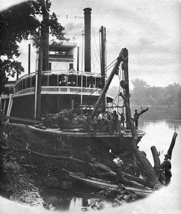 A steamboat using a crane to remove logs from a riverbank, with people working on the boat.