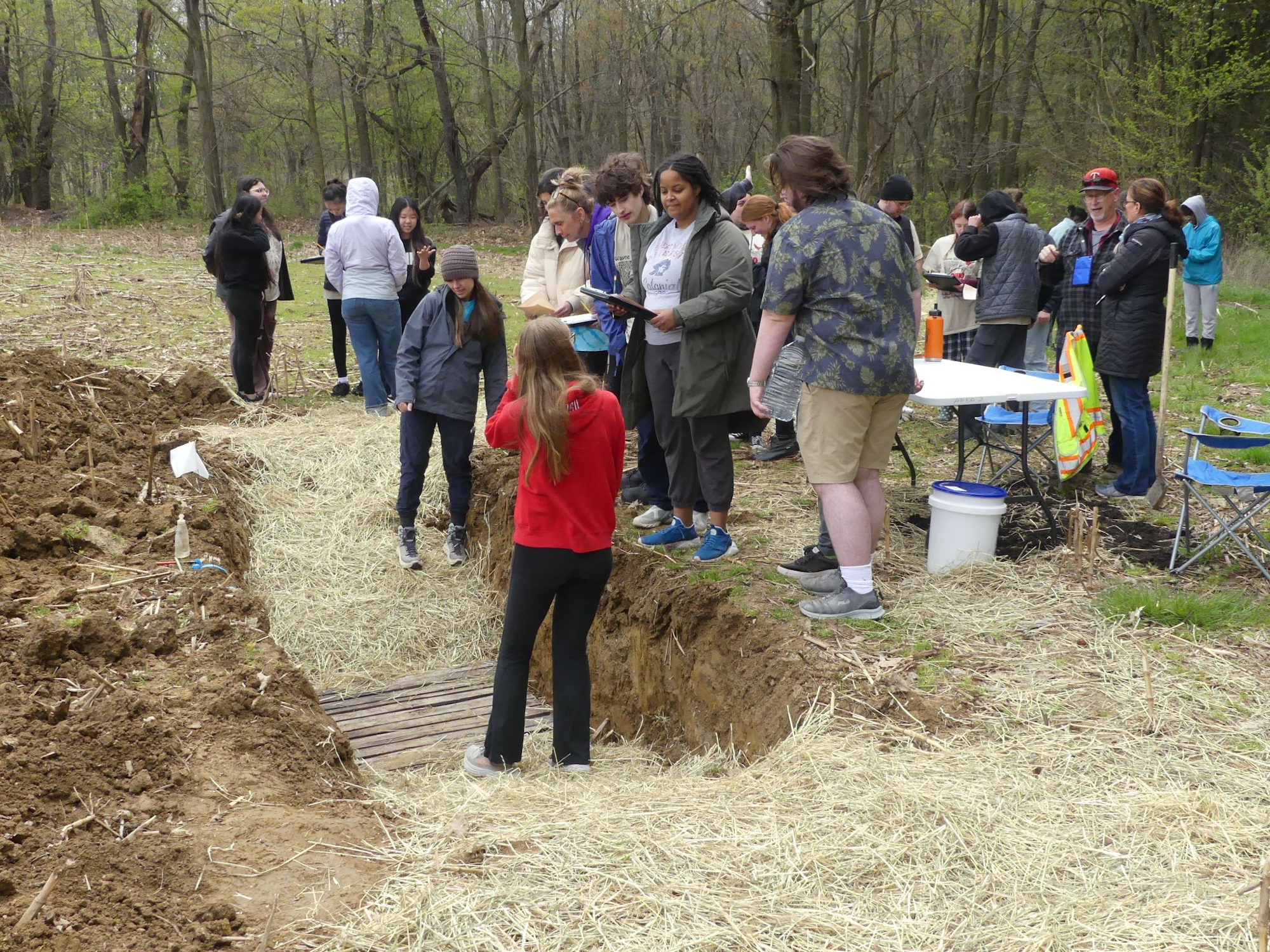 A group of people stands around a trench in a field, some taking notes, with a table and chairs nearby.