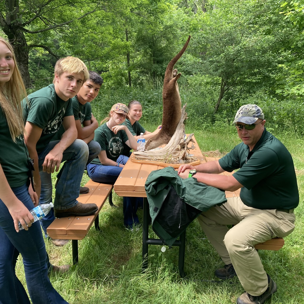 A group of people seated at a picnic table in a forest setting, looking at a taxidermied animal on the table.