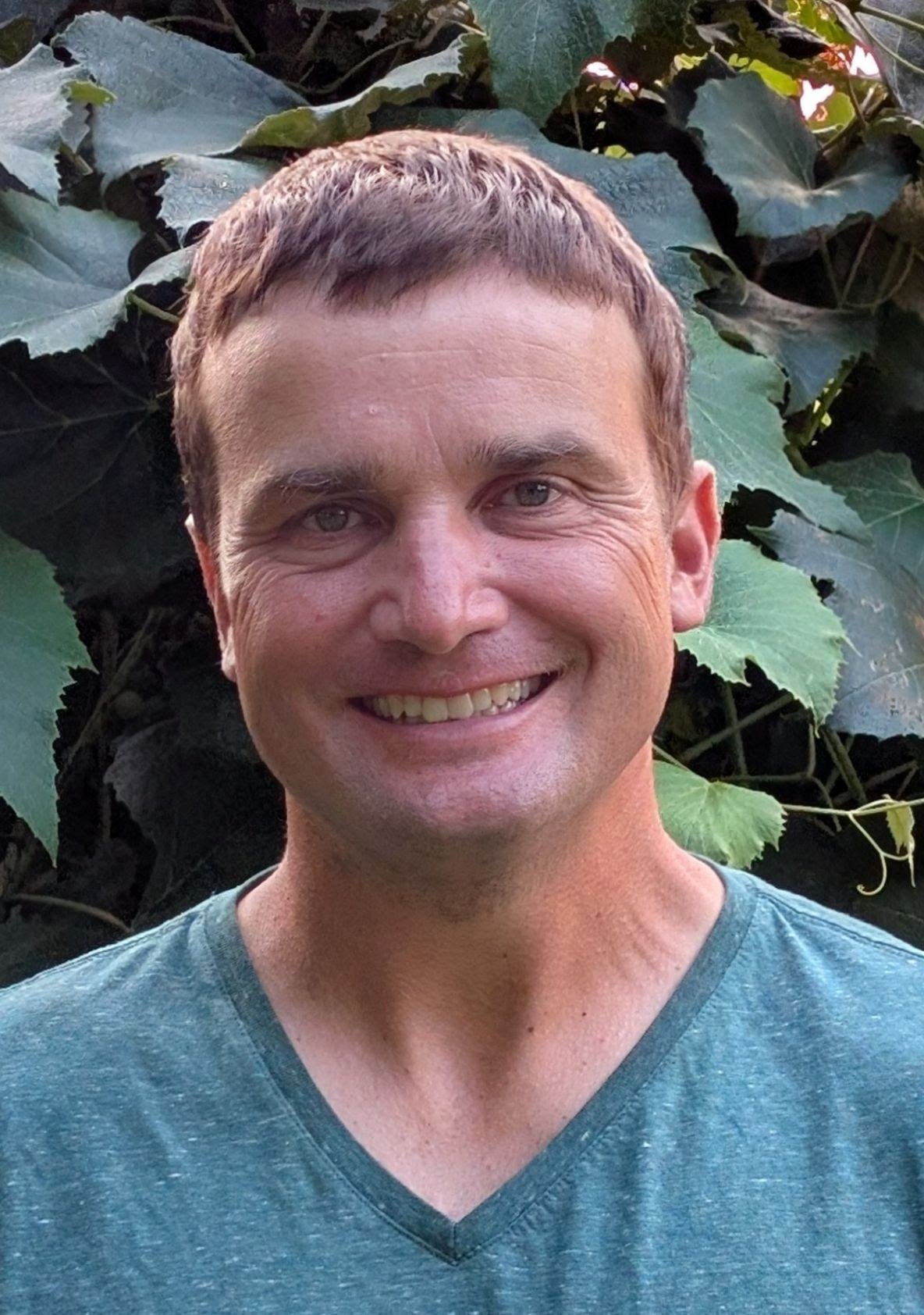 A person smiling, wearing a green shirt, with green leaves in the background.