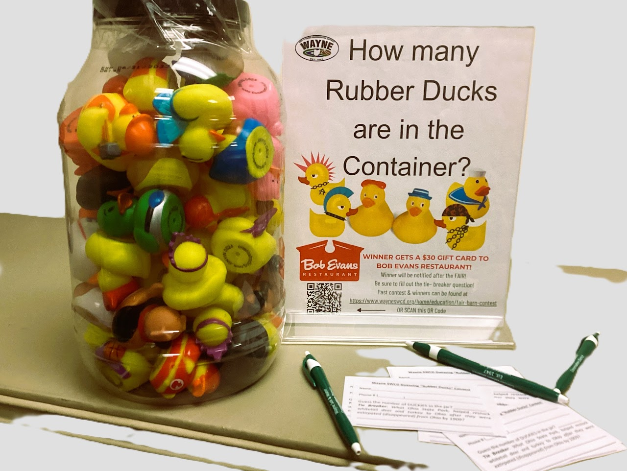 A jar filled with rubber ducks next to a sign asking, "How many Rubber Ducks are in the Container?" Pens and slips are in front.