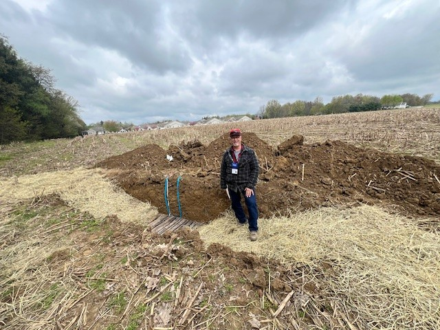A person stands near a large dug-up dirt area in a field, with cloudy skies above.