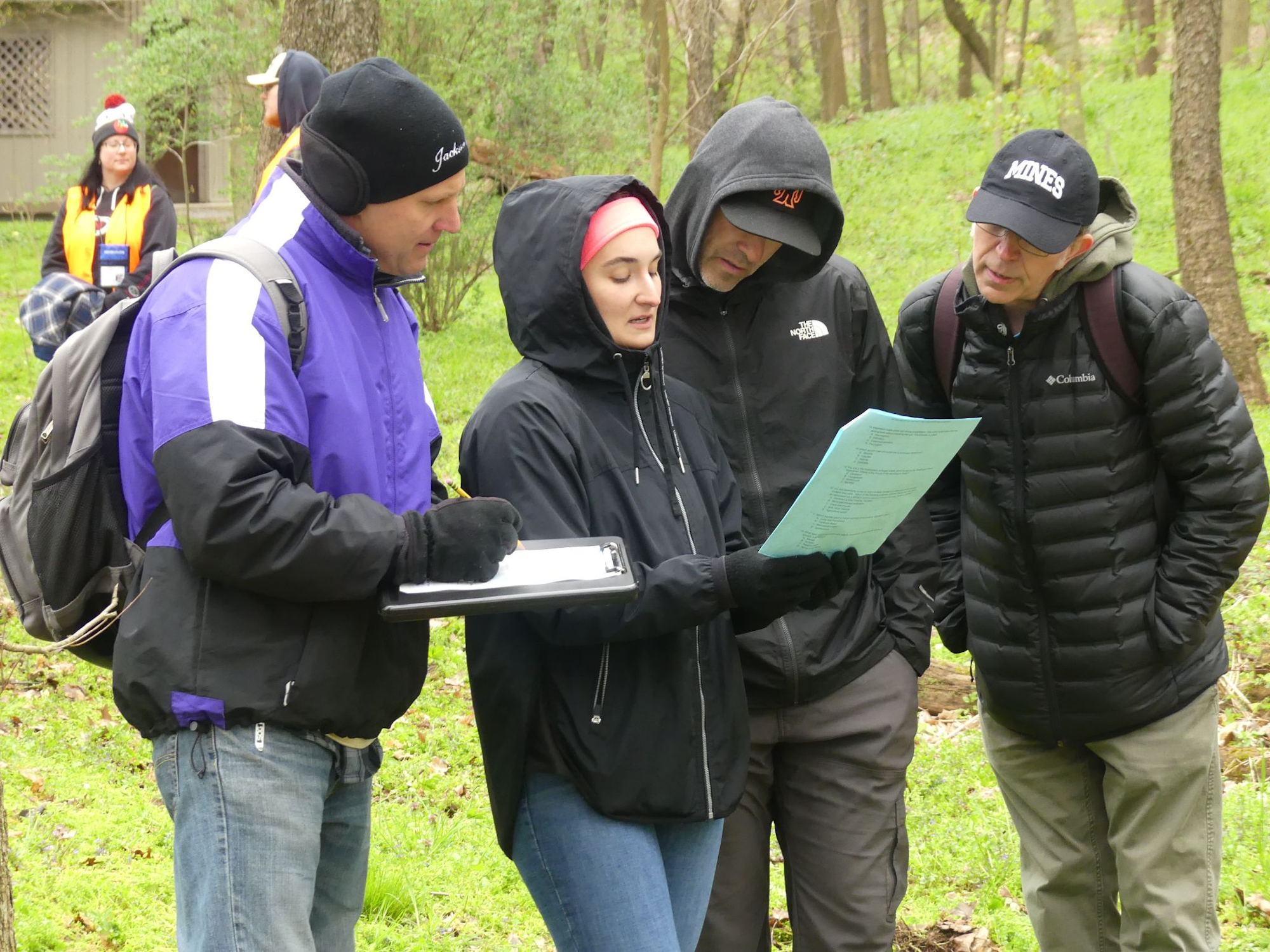 A group of people in outdoor clothing are examining a document in a wooded area.