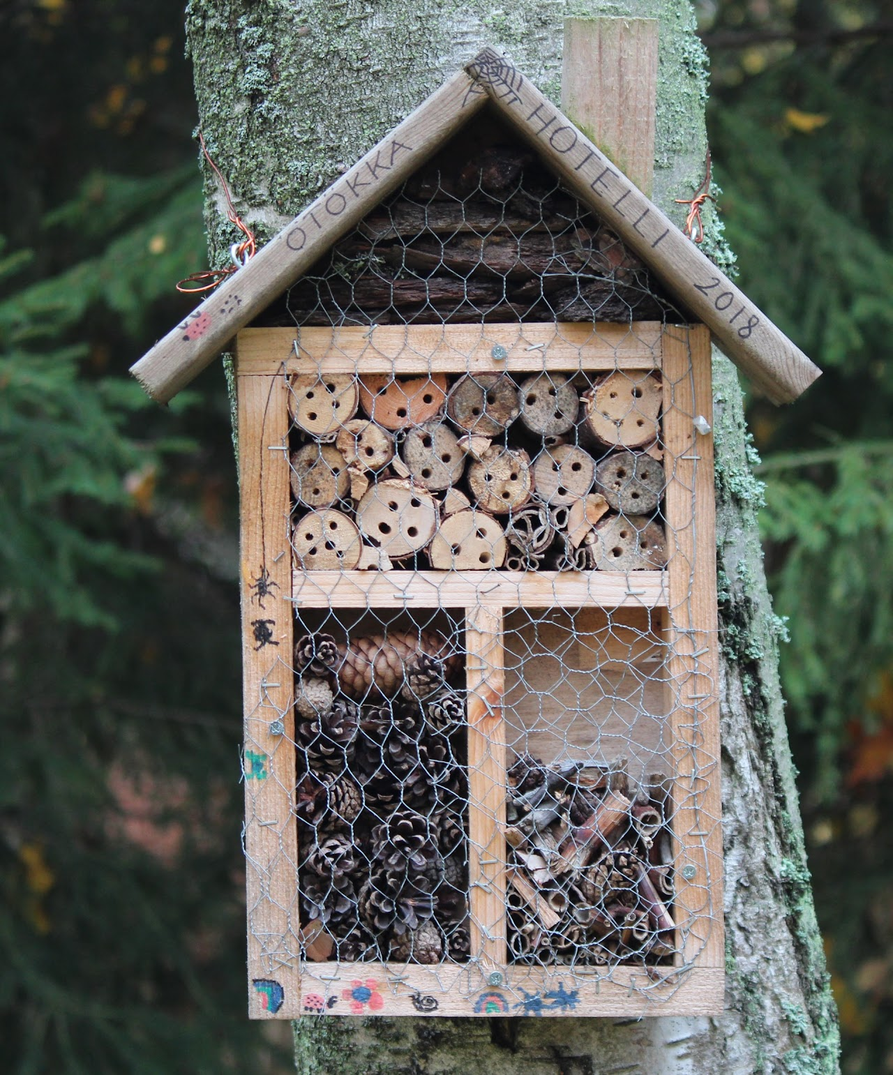 Wooden insect hotel filled with logs and pinecones, mounted on a tree.