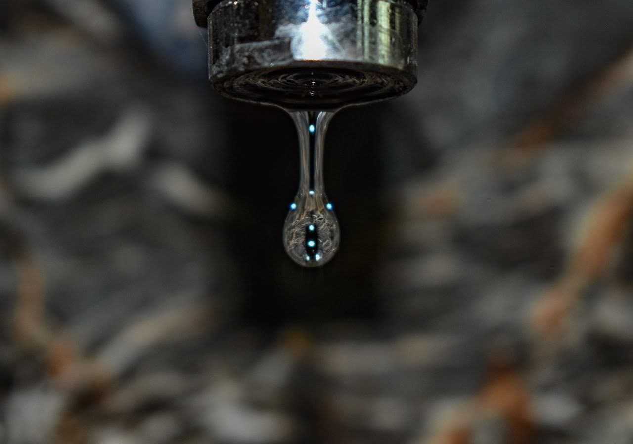 A close-up of a water droplet hanging from a faucet, with blurred background.