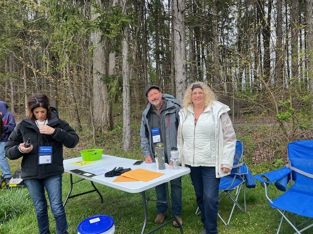 Three people at an outdoor table with papers and a green bowl, surrounded by trees.