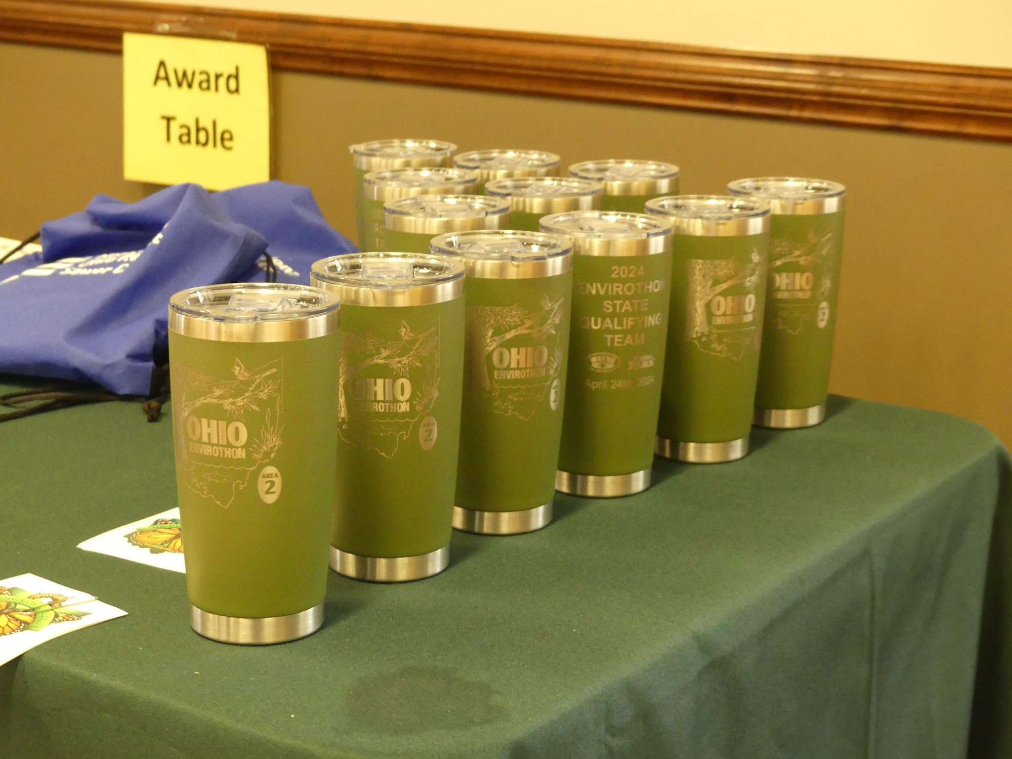 Green tumblers for Ohio Envirothon 2024 on a table labeled "Award Table," with blue bags in the background.