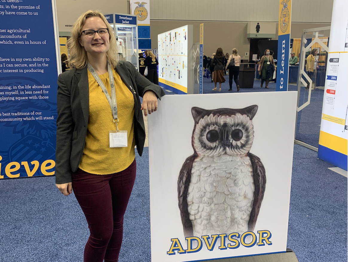 Person posing next to an "Advisor" owl poster at an indoor event with a lanyard.