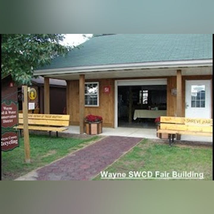 A wooden building with a green roof, open door, benches, plants, and a sign for Wayne SWCD Fair Building.