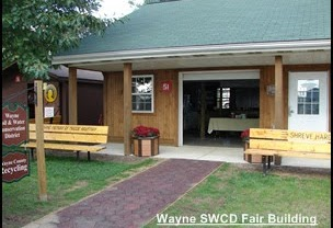 A wooden building with a green roof, open door, benches, plants, and a sign for Wayne SWCD Fair Building.