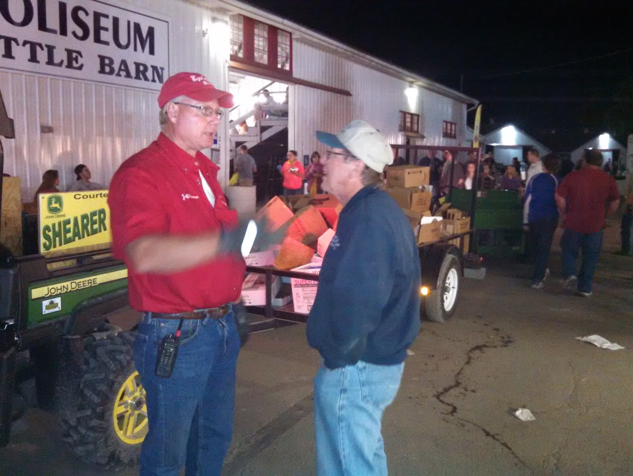 Two men talking outside a barn with a crowd in the background, near John Deere equipment.
