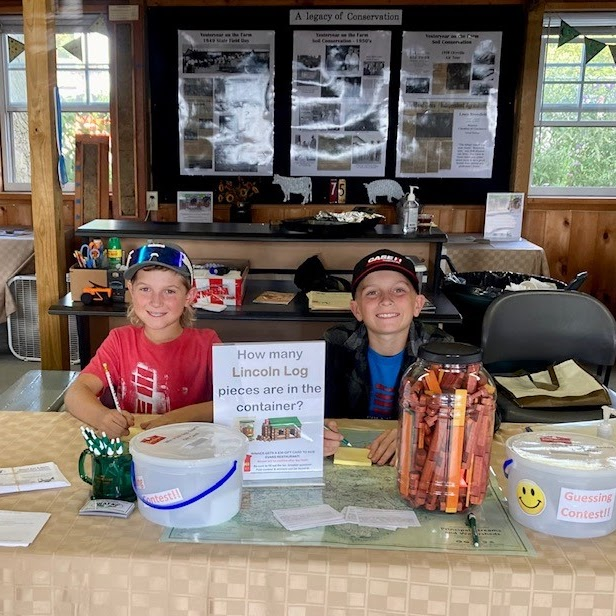 Two smiling kids behind a table with a Lincoln Log guessing contest and a jar filled with Lincoln Logs.