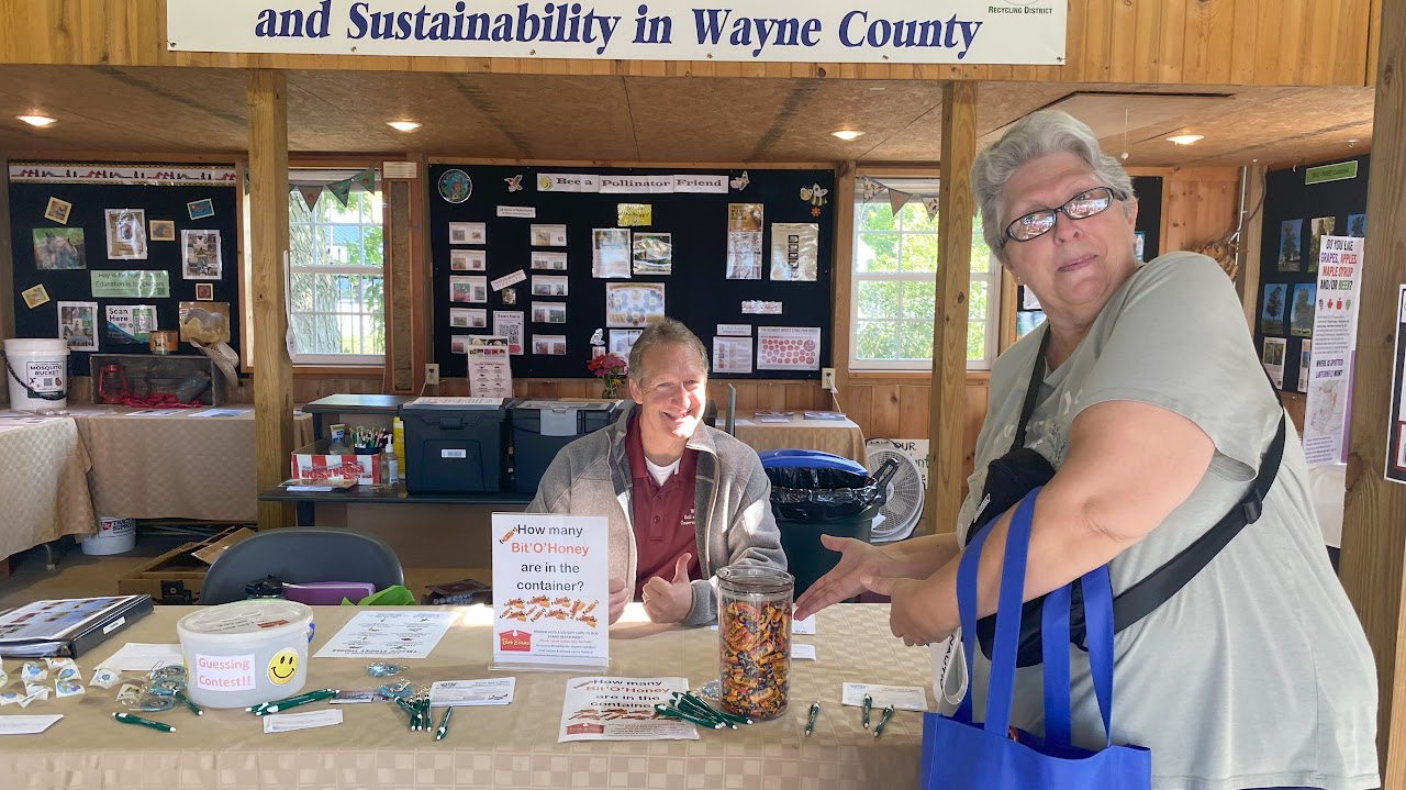 Two people at a table with a candy guessing contest, under a sign about recycling and sustainability in Wayne County.