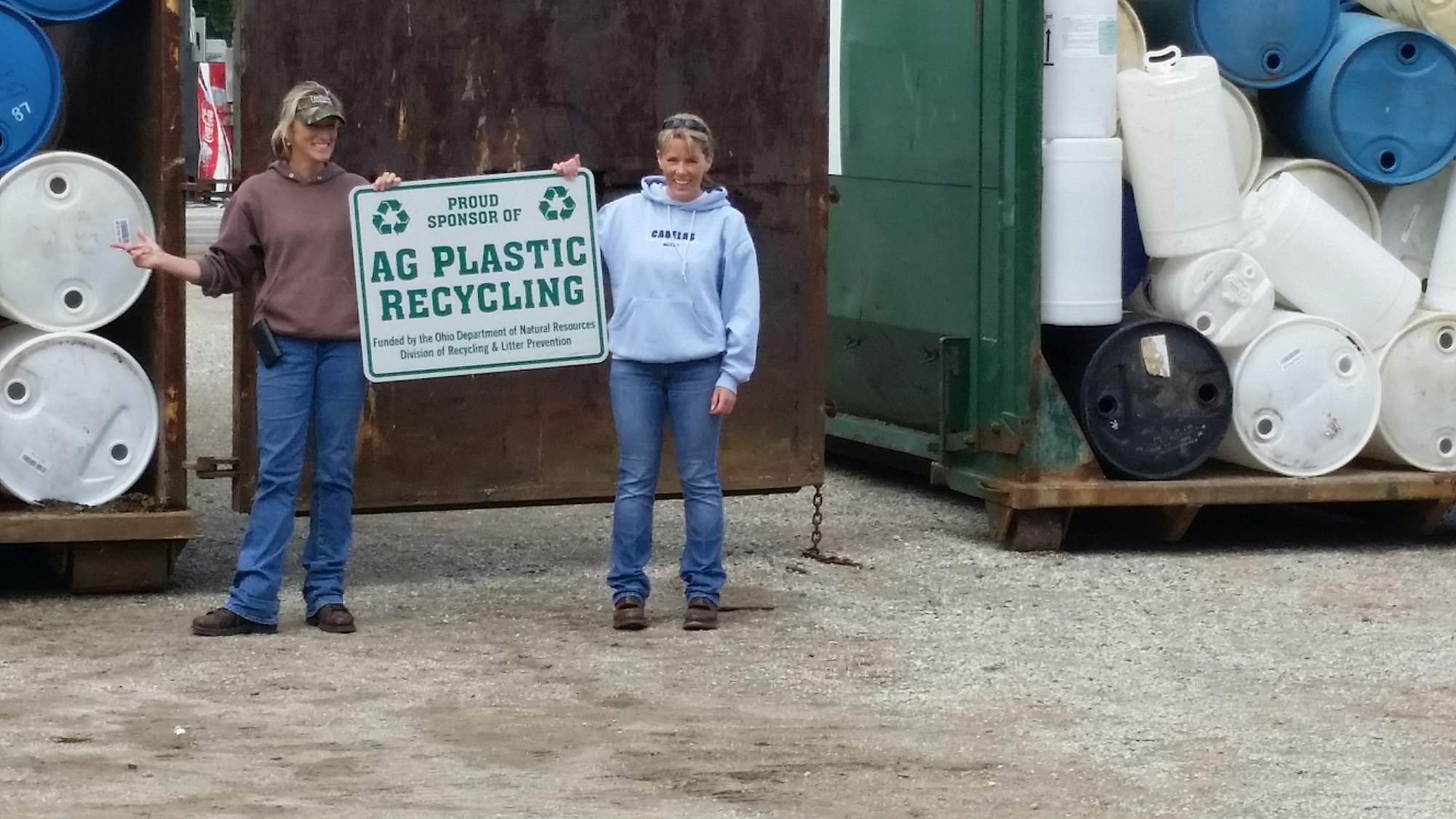 Two women stand by a sign promoting agricultural plastic recycling, with barrels stacked behind them.