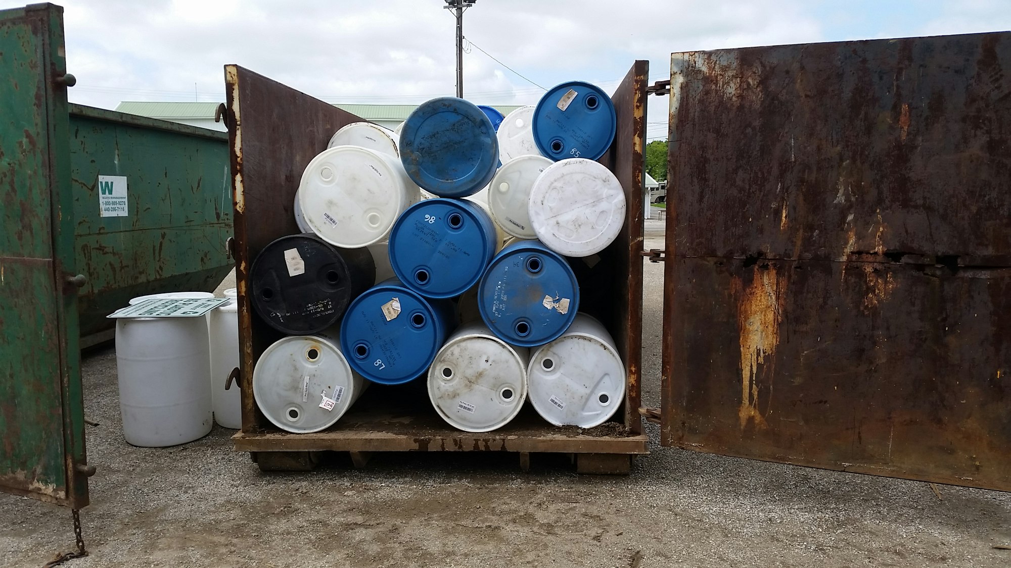 The image shows a dumpster containing stacked blue and white barrels, with some labels visible.