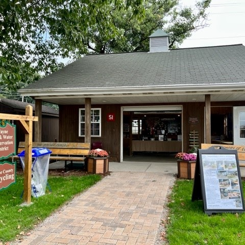 Small building with open entrance, benches, a sign, and recycling bin in front; surrounded by trees and greenery.