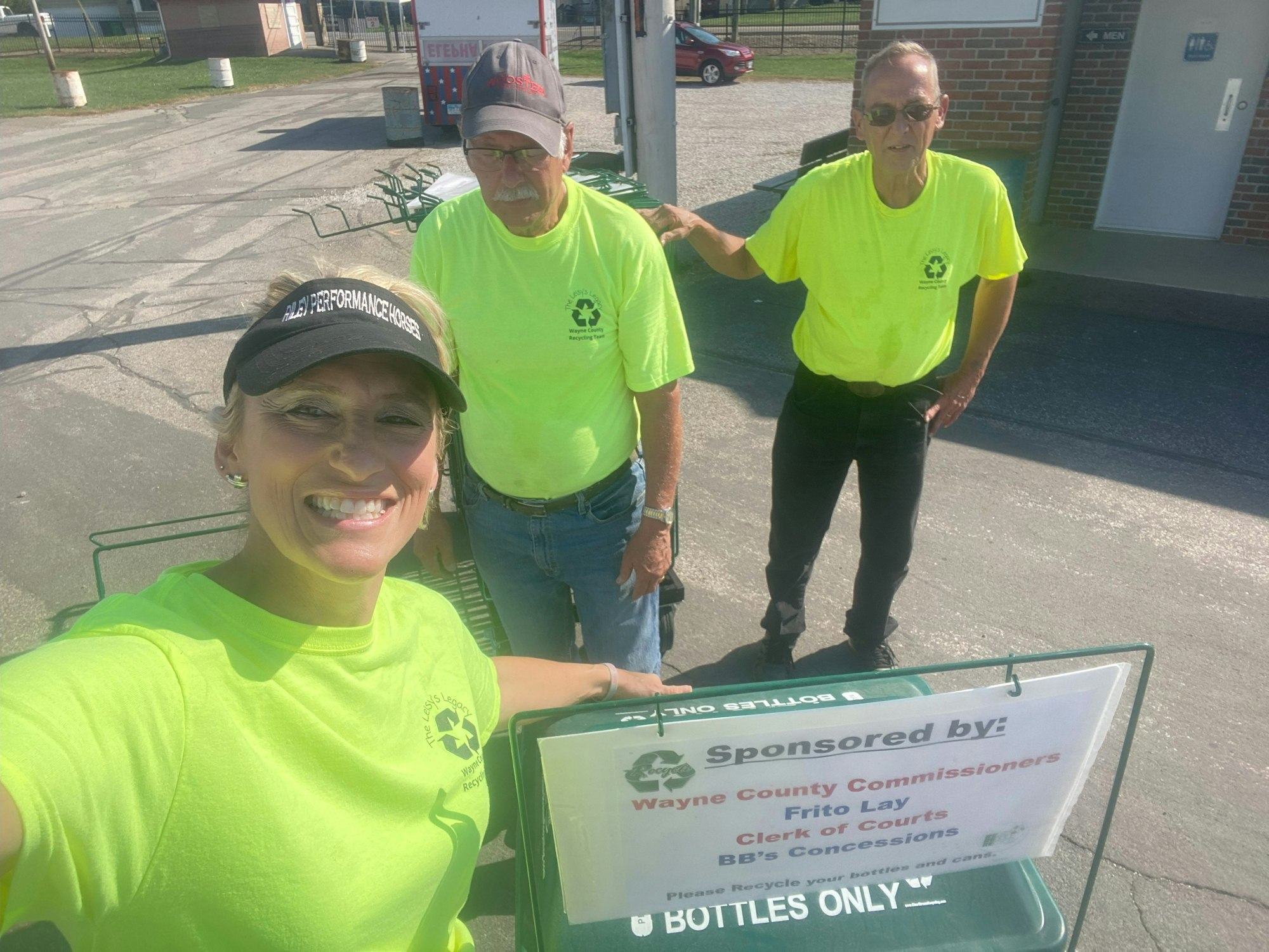 A group of three people in bright shirts near recycling bins, promoting a recycling initiative sponsored by local organizations.