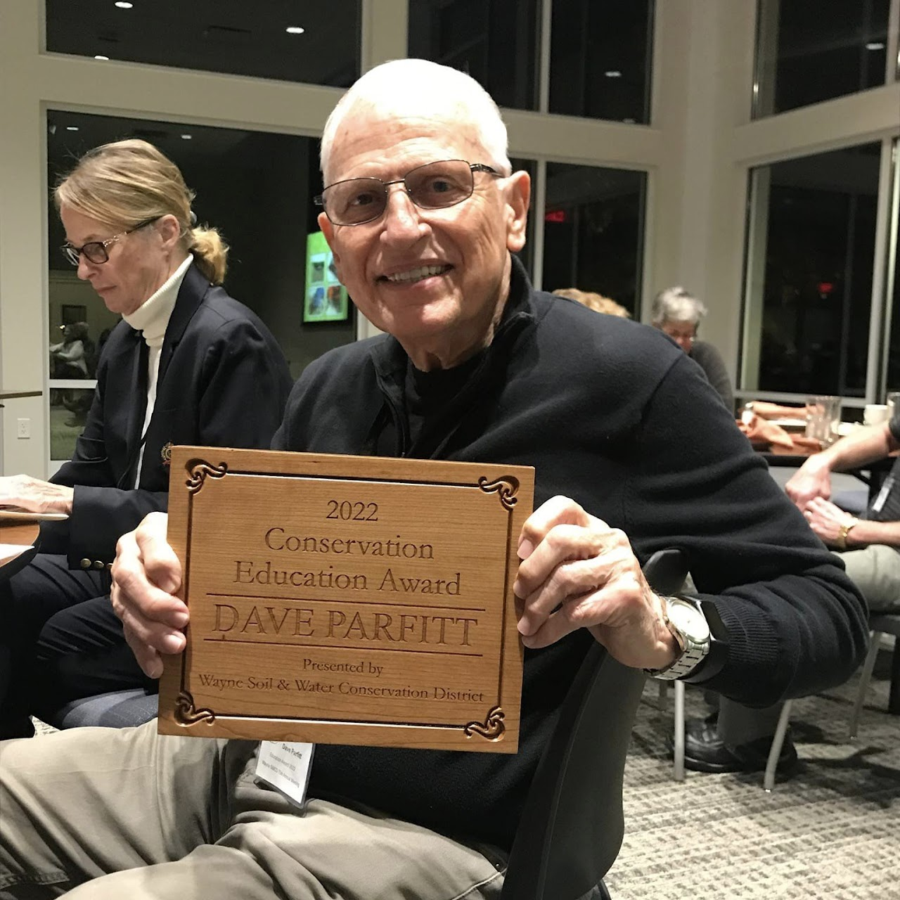 A person holding a "2022 Conservation Education Award" plaque at an event, others are seated around.