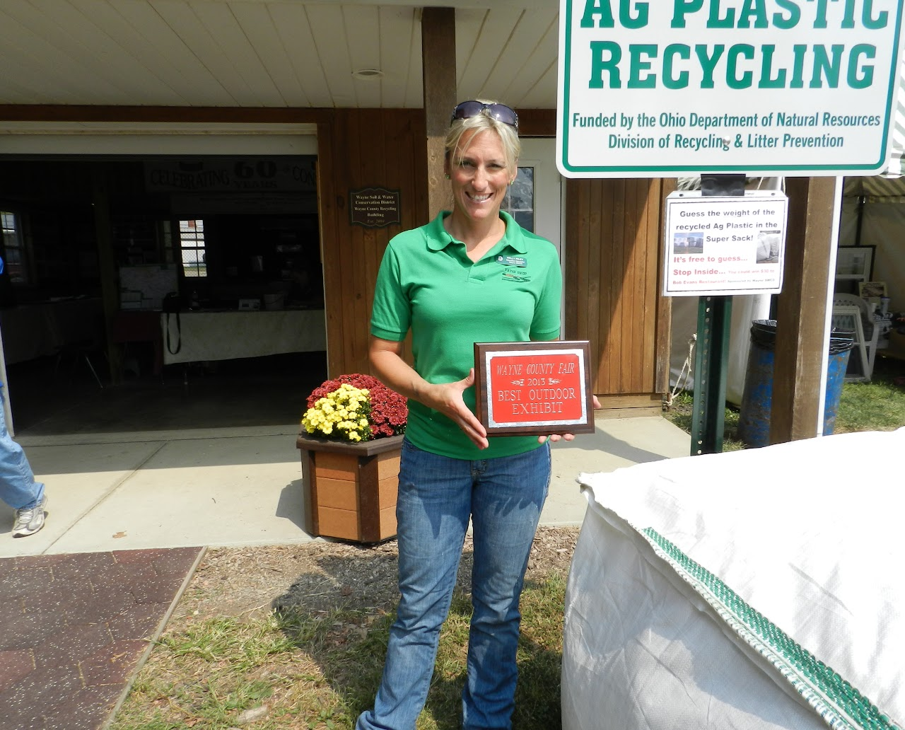 Person holding a plaque for "Best Outdoor Exhibit" at the Wayne County Fair, standing by an "Ag Plastic Recycling" sign.