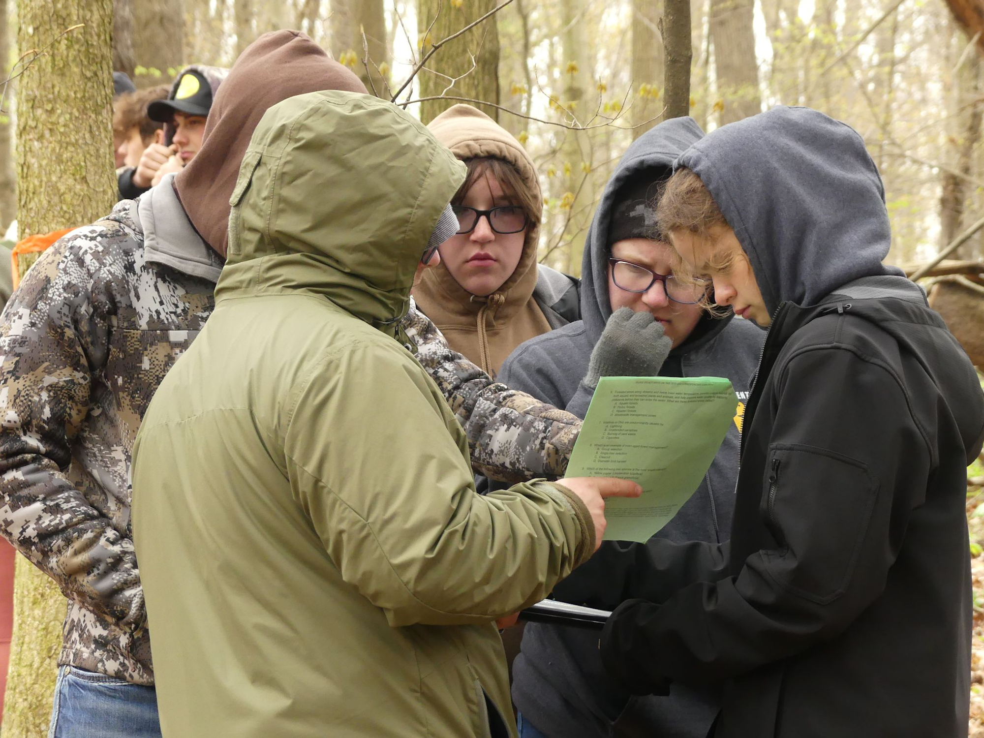 A group of people in hoodies and jackets are gathered in a wooded area, looking at a paper document.