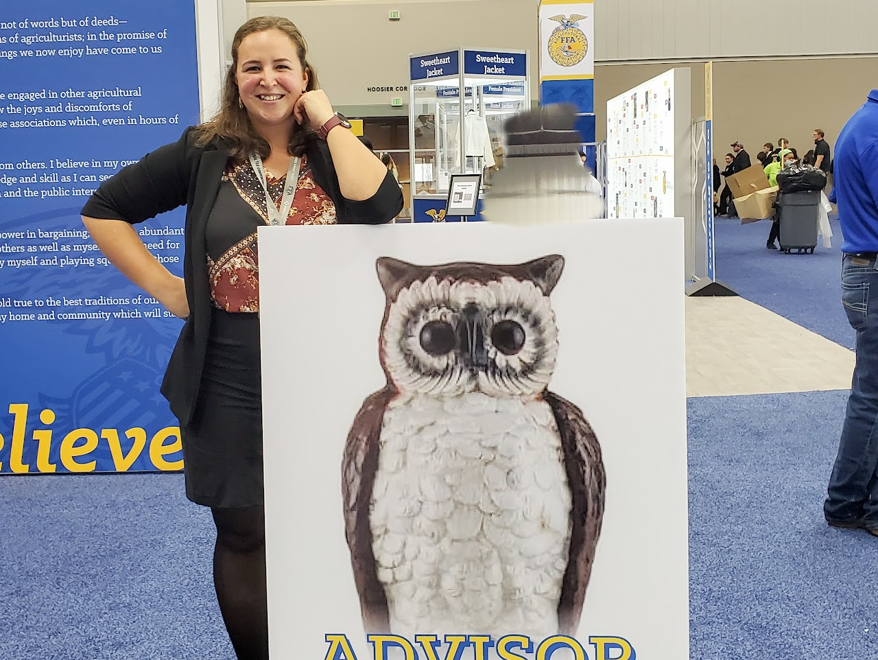 A person stands beside a sign with an owl illustration labeled "ADVISOR." There are people and booths in the background.