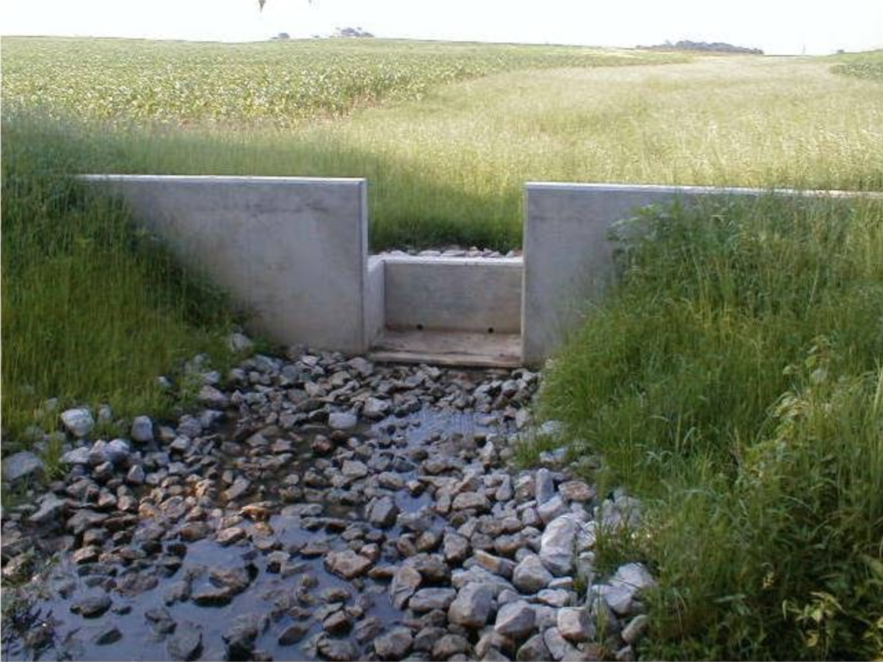 A stone-lined drainage ditch with water, enclosed by a concrete structure, surrounded by grassy fields.