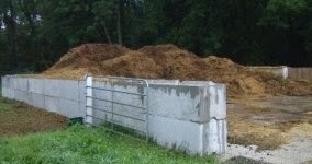 A large pile of compost or manure is enclosed by concrete walls and a metal gate, situated outdoors.