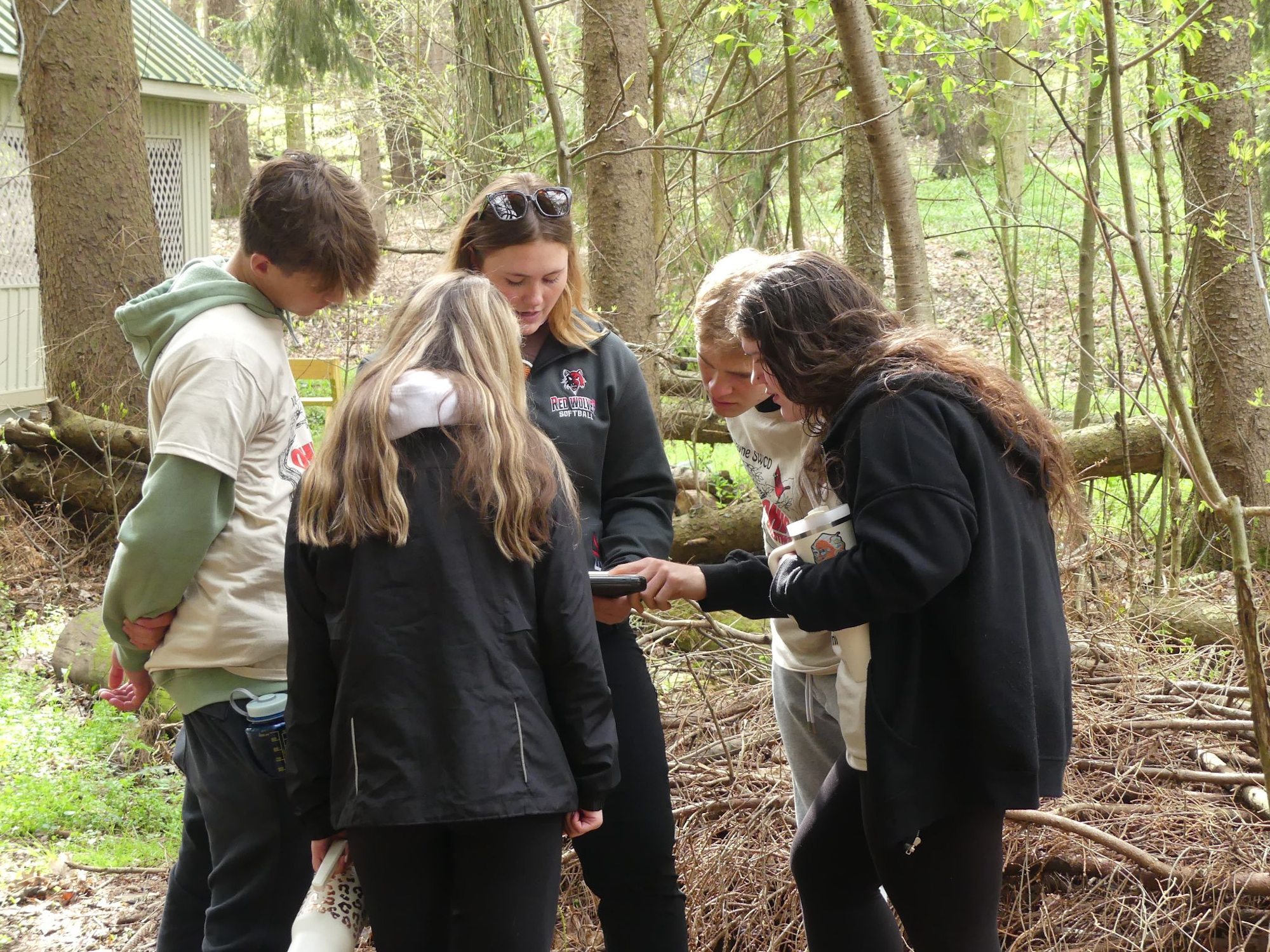 A group of people in a wooded area, gathered around a device, possibly a phone, engaged in a shared activity.