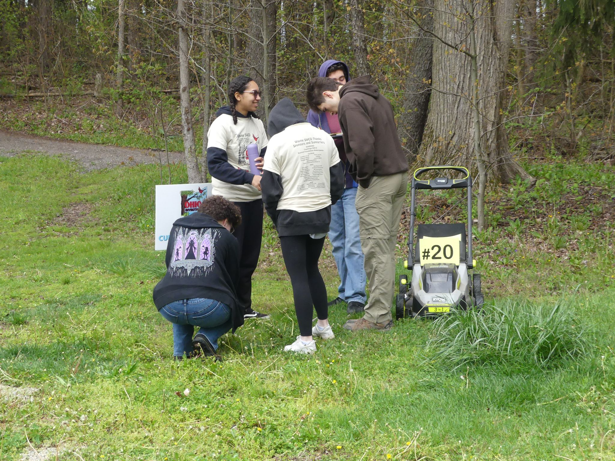 Group of people gathered around a lawnmower labeled #20, outdoors with trees in the background.