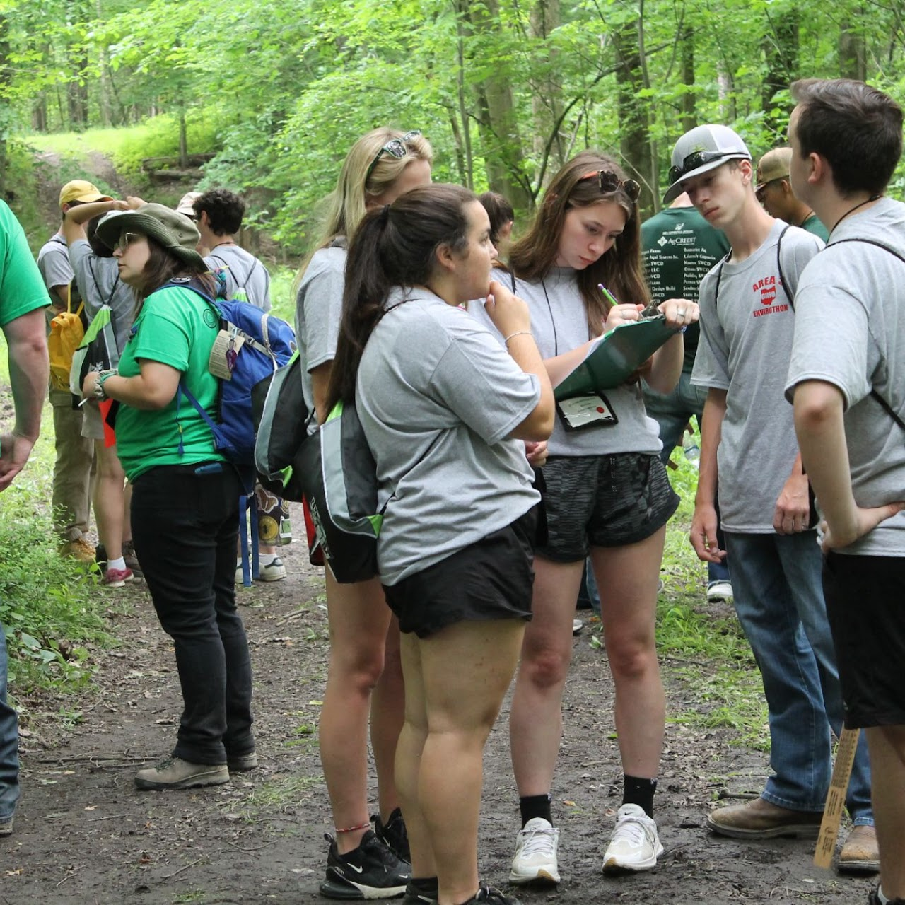 A group of people outdoors in a forest, wearing casual clothes, and looking at a clipboard.