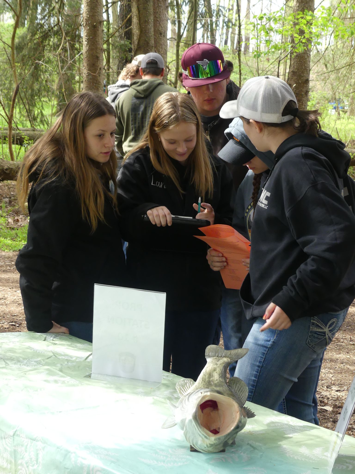 A group of people is gathered in a forest setting, examining papers and a large fish model on a table.