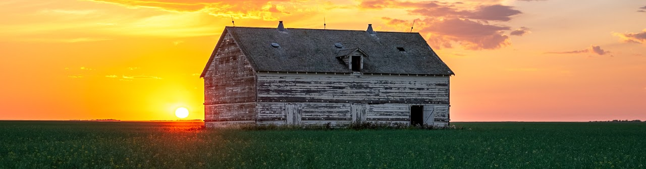 A weathered wooden barn in a green field at sunset, with a vibrant orange sky.