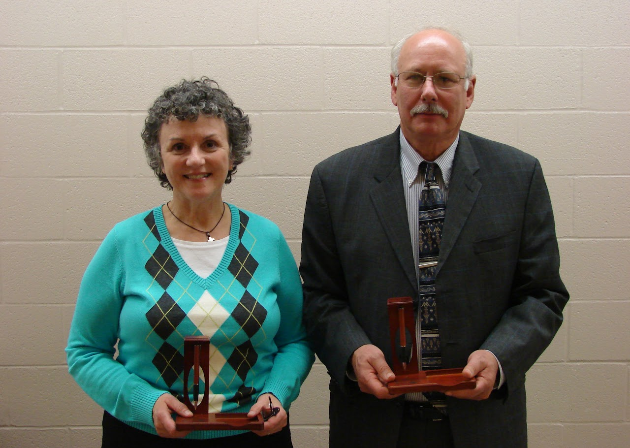 Two people standing with awards, one is wearing a turquoise sweater, and the other is in a suit and tie.