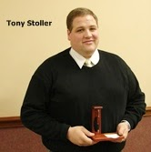 A person holding a small wooden object, wearing a black sweater over a white shirt, standing indoors with a beige background.