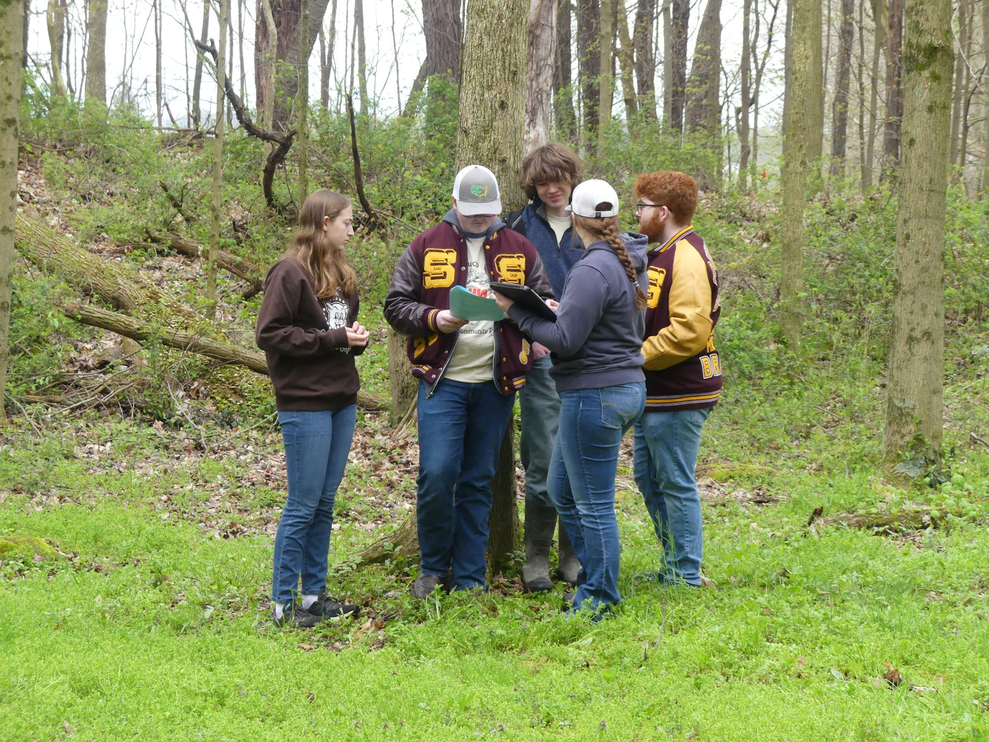 Five people in a forest, some wearing letterman jackets, looking at papers.