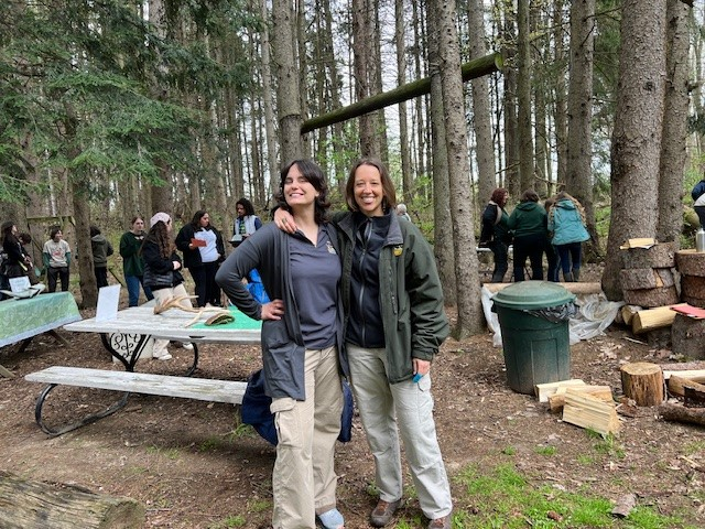 Two people smiling in a wooded area with a group of people in the background, near a picnic table and logs.