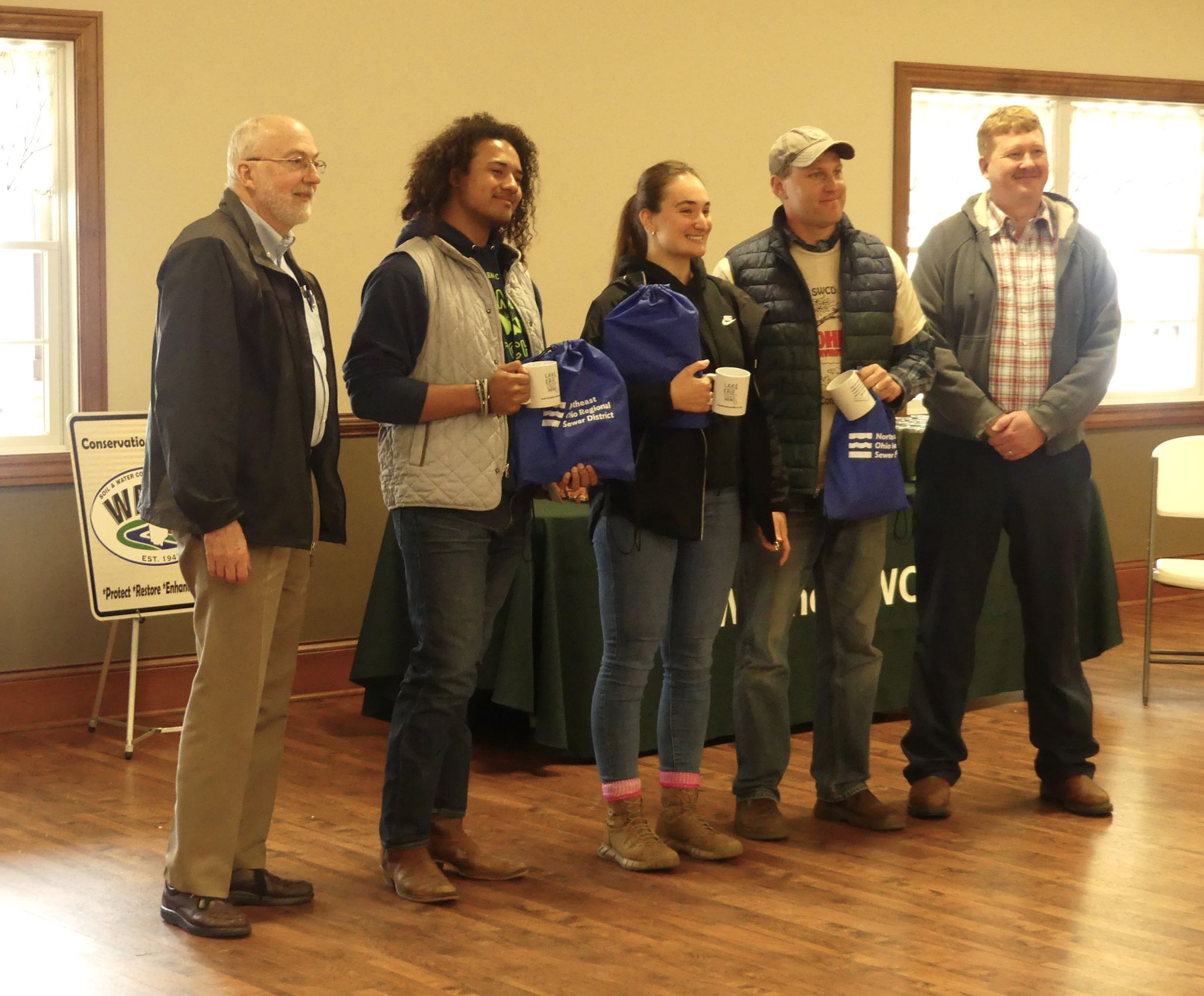 A group of people posing with mugs and blue bags at an indoor event; a conservation sign is visible in the background.