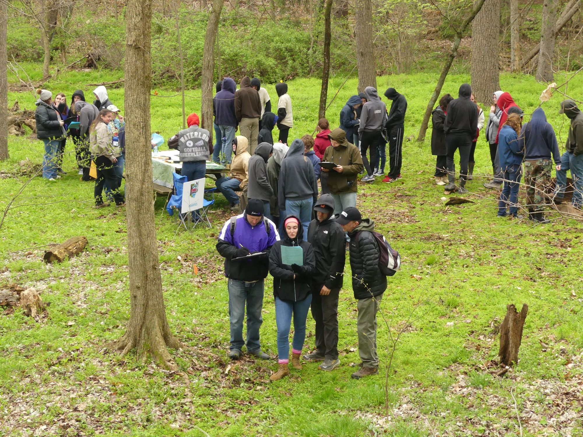 A group of people dressed warmly gather in a wooded area with tables, seemingly for an outdoor event or activity.