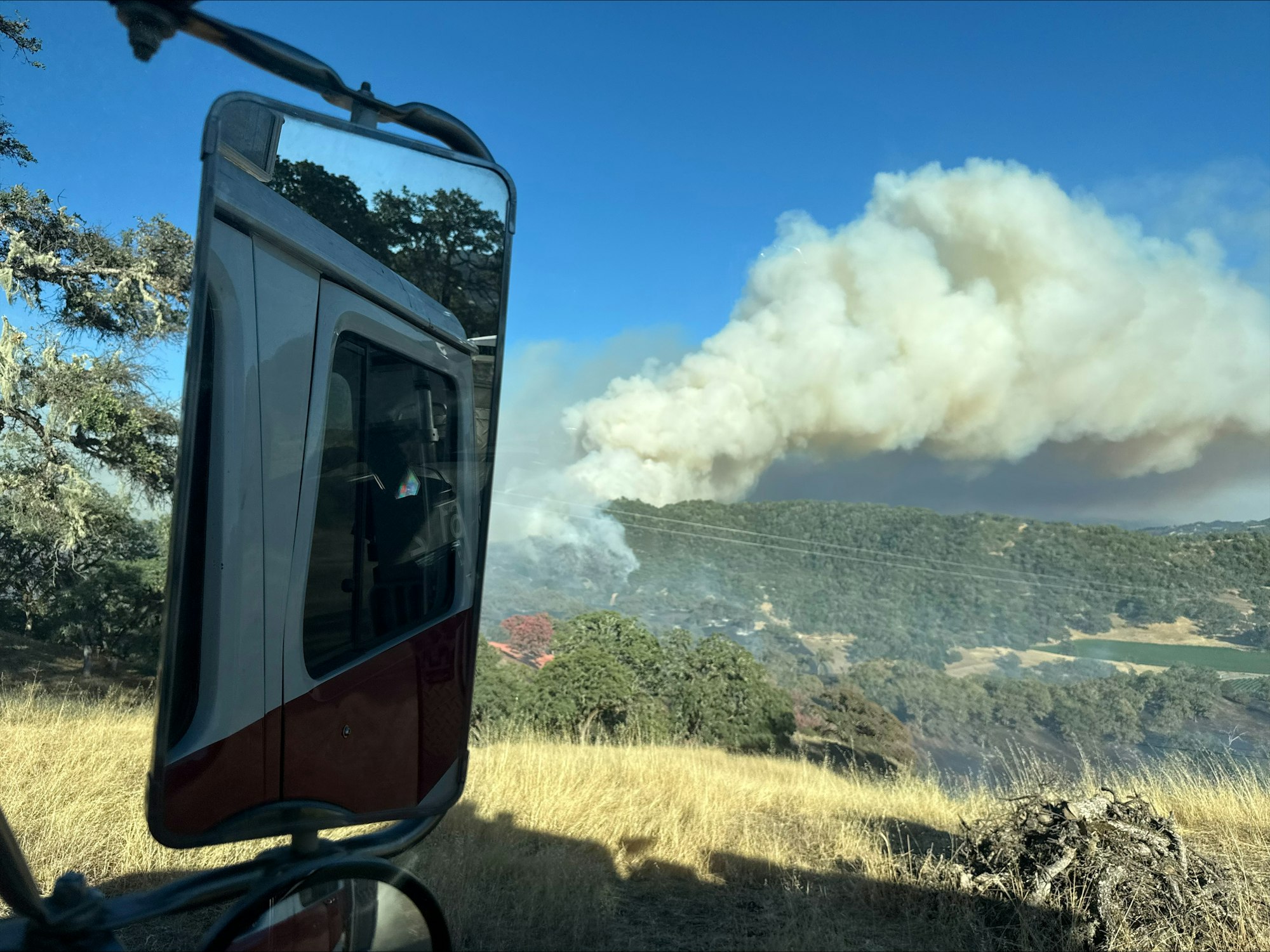 A view from a vehicle showing smoke rising from a wildfire on a hillside, with dry grass and trees nearby.