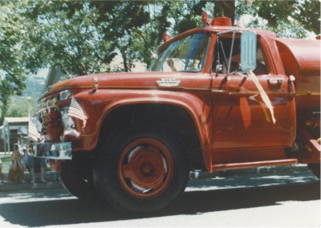 A vintage red fire truck with ribbons on the street, surrounded by trees.
