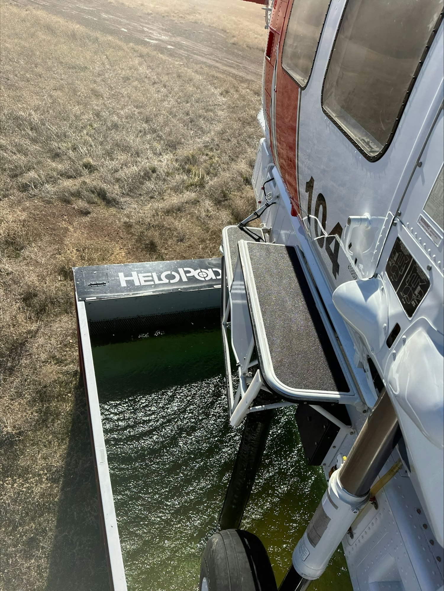 A helicopter's open door facing a grassy field, revealing the step and landing skid with water below.