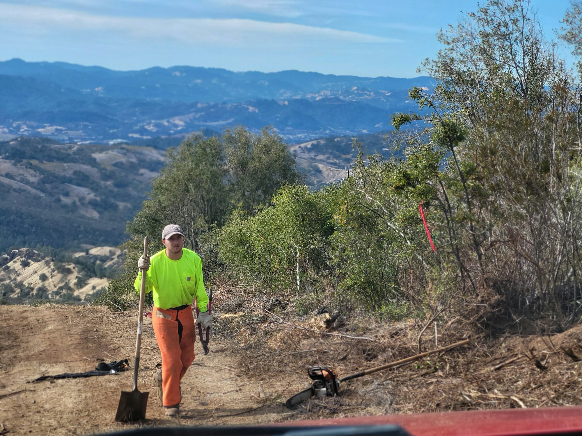 A person in bright work clothes carries a shovel and tool on a dirt path with a mountainous landscape in the background.