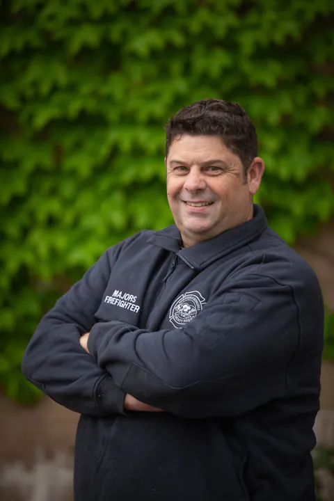 A smiling firefighter in a dark uniform stands with arms crossed in front of a green leafy background.