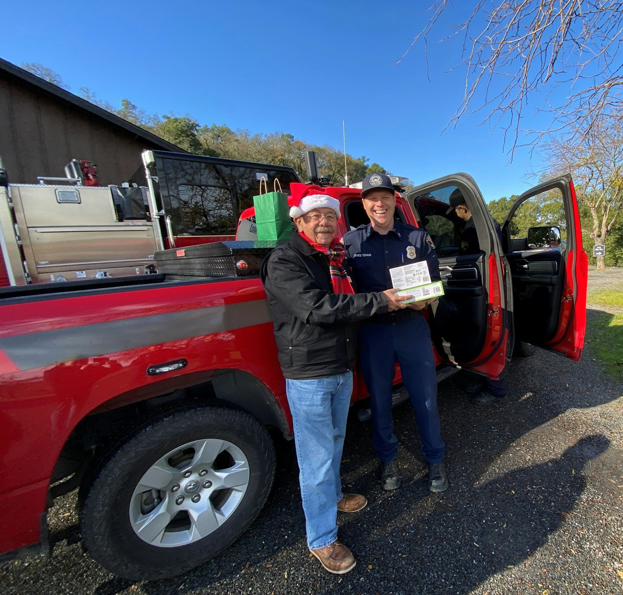 Two people smiling near a red truck, one wearing a Santa hat, holding a gift.