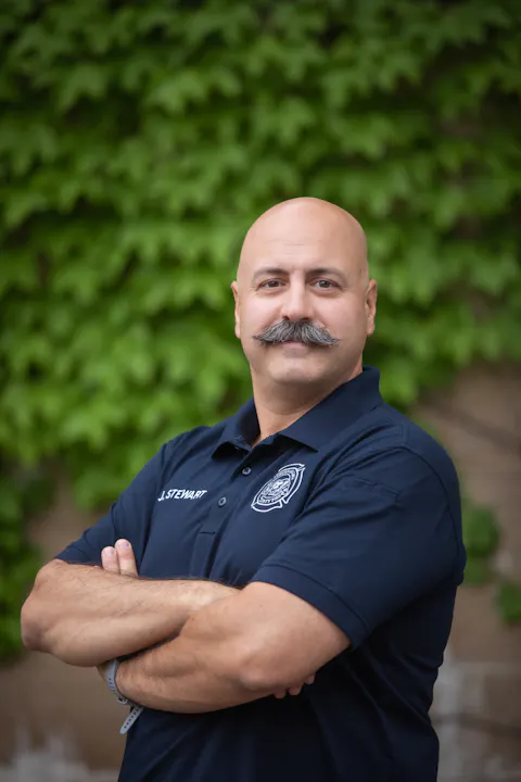 A bald man with a mustache in a navy polo shirt stands confidently against a green leafy background, arms crossed.
