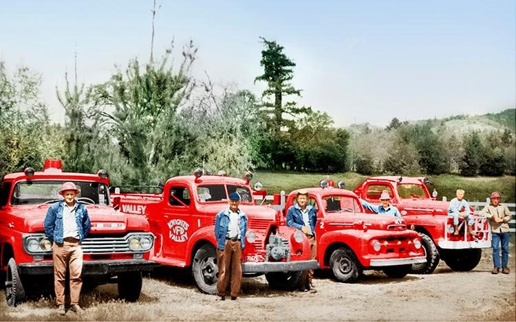 Five vintage red fire trucks lined up, with six people standing in front, in a rural outdoor setting.