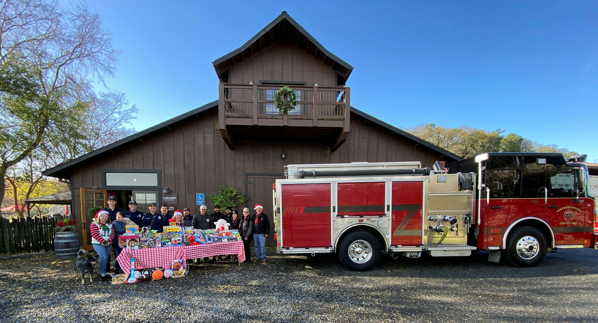 A group of people stand by a red fire truck with a table of toys in front of a barn-like building.